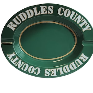 May include: An oval-shaped dark green ashtray featuring the text "RUDDLES COUNTY" in white capital letters along the rim. The ashtray has a smooth, glossy finish with a thin gold-colored border accenting the inner basin and two cigarette rests.