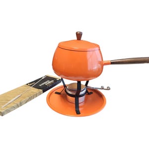 May include: An orange fondue pot with a wooden handle and lid, set on a matching orange stand with a black metal frame. A box of fondue forks is to the left. The pot is designed for cooking and serving.