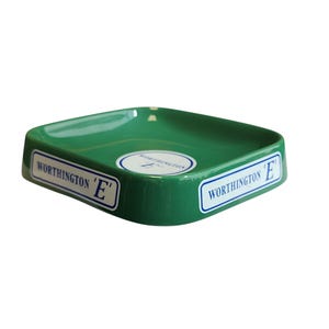 Può includere: Un posacenere quadrato in ceramica verde con angoli arrotondati. I lati esterni e il centro della vaschetta presentano etichette rettangolari bianche con bordi blu, ciascuna con il testo WORTHINGTON 'E' in un carattere serif blu scuro.