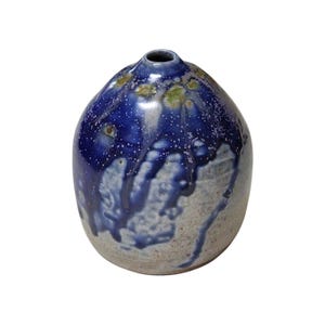 Może przedstawiać: Ręcznie wykonany ceramiczny wazon o zaokrąglonym kształcie i małym otworze. Wazon ma nakrapianą niebieską glazurę, która spływa po jaśniejszej, teksturowanej podstawie. Góra ma ciemnoniebieską glazurę z małymi, jasnymi kropkami.