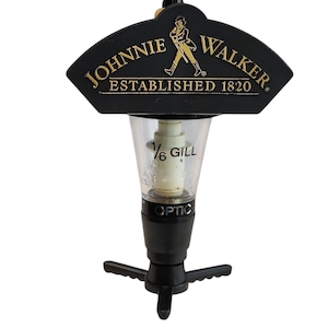 Può includere: Un dosatore per liquori nero Johnnie Walker con logo dorato e la scritta Established 1820. La camera di misurazione in plastica trasparente riporta le diciture 1/6 GILL e OPTIC, con un meccanismo di erogazione nero a tre punte alla base.