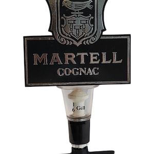 Può includere: Un dosatore per bottiglia di cognac Martell con insegna rettangolare nera, scritte argentate e stemma con uccello. Il componente di misurazione in plastica trasparente riporta la scritta 1/6 Gill ed è fissato a una base nera con leva.