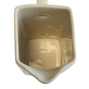 May include: A cream ceramic creamer with a rectangular body and a spout. The creamer has a small handle on top and a slightly textured surface. The interior is a light beige colour.