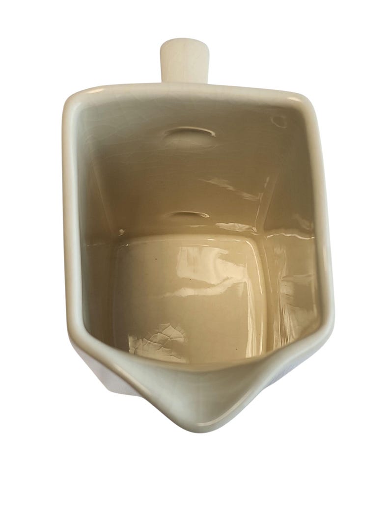 May include: A cream ceramic creamer with a rectangular body and a spout. The creamer has a small handle on top and a slightly textured surface. The interior is a light beige colour.