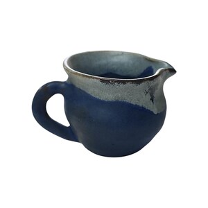 May include: A handmade ceramic creamer with a dark blue base and a light blue rim. The creamer has a curved handle and a spout for easy pouring. The design is simple and elegant, suitable for serving cream or milk.