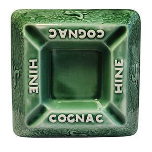 Può includere: Un posacenere quadrato in ceramica verde con le parole "COGNAC" e "HINE" in lettere bianche. Il posacenere ha un centro incassato e quattro posa-sigarette. La superficie presenta un motivo strutturato a forma di foglia.
