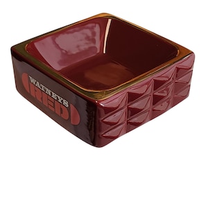 Può includere: Una ciotola quadrata in ceramica dai bordi profondi con una finitura rosso scuro lucida. L'esterno presenta un motivo geometrico strutturato di triangoli in rilievo su due lati e la scritta "WATNEYS RED" stampata in bianco e rosso sul davanti.