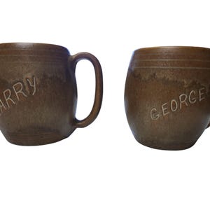 以下が含まれることがあります： 取っ手付きの2つの茶色のセラミックマグ。それぞれに名前が刻まれています。1つのマグには「LARRY」、もう1つには「GEORGE」と書かれています。マグカップは素朴で手作りの外観をしています。