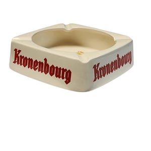 Può includere: Un posacenere quadrato in ceramica color bianco sporco con il testo rosso Kronenbourg stampato su due lati. Il posacenere ha una finitura liscia e lucida con quattro tacche distinte lungo il bordo superiore e una vaschetta centrale.