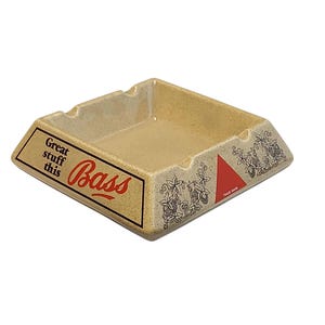 Può includere: Un posacenere quadrato in ceramica beige maculata con la scritta "Great stuff this" e "Bass" in lettere rosse e nere. Il design include un logo a triangolo rosso e disegni a linee nere di viti di luppolo e foglie sui pannelli laterali.