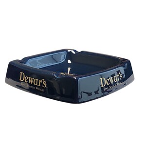 Può includere: Un posacenere quadrato in ceramica blu scuro con angoli arrotondati e quattro poggia sigarette. I lati presentano il testo dorato "Dewar's Fine Scotch Whisky" stampato su due facce adiacenti. L'interno è liscio, lucido e di colore blu profondo.
