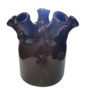 May include: A dark brown ceramic jug with a unique design. It features a cylindrical base and a top with four spouts. The jug has a glossy finish, and the spouts are angled outwards. This decorative piece is suitable for home decor.