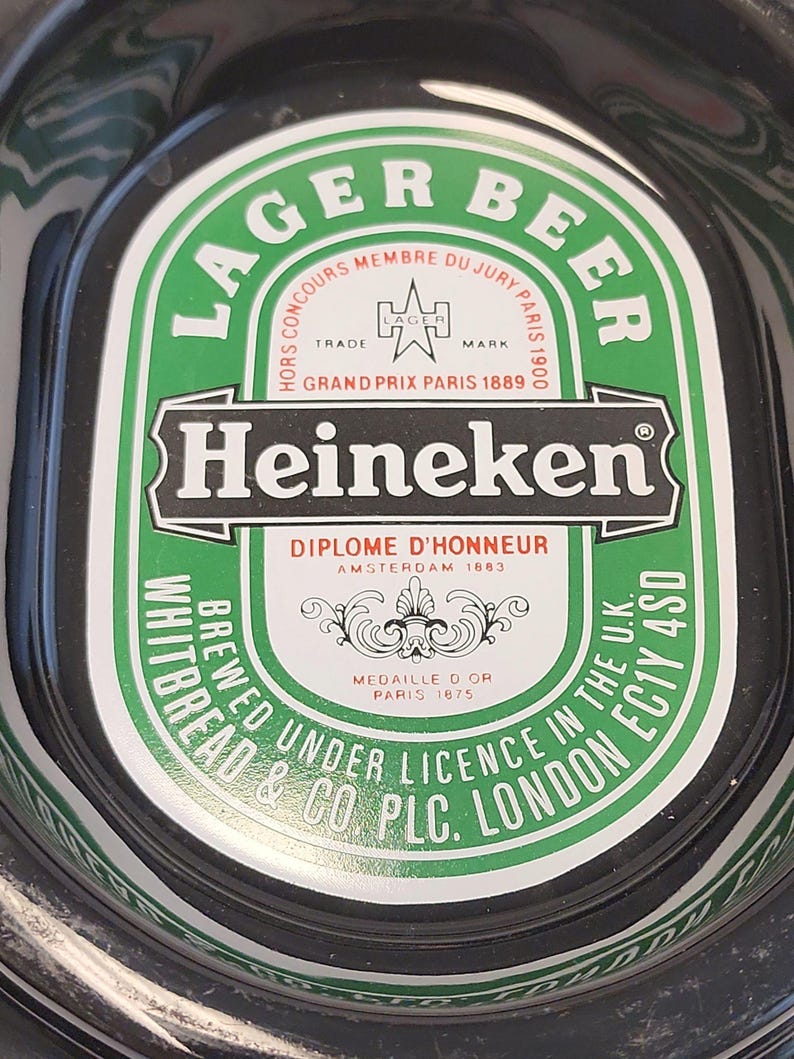 Puede incluir: Una bandeja ovalada negra y verde de cerveza Heineken con el texto LAGER BEER, Heineken, DIPLOME D'HONNEUR AMSTERDAM 1883, HORS CONCOURS MEMBRE DU JURY PARIS 1900, GRAND PRIX PARIS 1889, MEDAILLE D OR PARIS 1875 y WHITBREAD & CO PLC LONDON EC1Y 4SD.