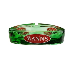 Può includere: Un posacenere rotondo in vetro verde traslucido caratterizzato da un logo ovale rosso con il testo bianco MANNS stampato sul lato. Il posacenere presenta tre tacche distinte lungo il bordo superiore e una finitura liscia e lucida.