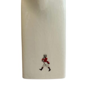 May include: An off-white ceramic flask with a rectangular shape and a small illustration of a person in a red coat and black boots. The flask has a smooth, glossy finish.