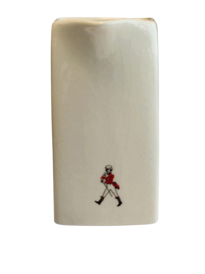 May include: An off-white ceramic flask with a rectangular shape and a small illustration of a person in a red coat and black boots. The flask has a smooth, glossy finish.