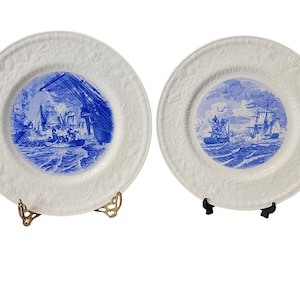 以下が含まれることがあります： 海に船が描かれた青と白のイラストが施された白いセラミック製のプレート2枚。プレートは縁が高く、装飾的な縁取りが施されています。