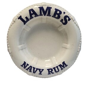 Può includere: Posacenere in ceramica bianca a forma di salvagente, con la scritta "LAMB'S NAVY RUM" in blu navy. Il posacenere ha una superficie screpolata e un bordo perlato.