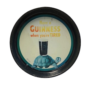 Può includere: Un vassoio da portata rotondo in metallo nero con una pubblicità vintage raffigurante una tartaruga blu che trasporta un bicchiere di birra scura. Il testo recita Have a GUINNESS when you're TIRED. Il design include un bordo circolare verde acqua e il nome Gilroy.
