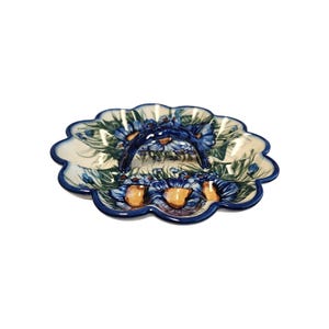 May include: A ceramic serving dish with a scalloped edge and a handle. The dish features a floral design with blue flowers, green leaves, and orange centers on a cream background. The handle and rim are dark blue.