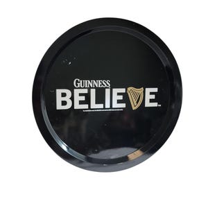 Può includere: Un vassoio da portata rotondo in metallo nero con la scritta bianca GUINNESS BELIEVE, in cui un'icona di arpa dorata sostituisce la lettera V. La superficie è lucida e riflettente, mostrando il logo del marchio e le informazioni sul marchio.