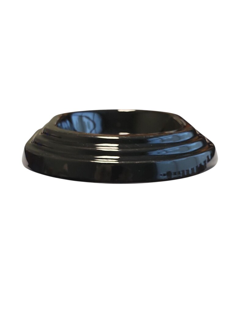 Puede incluir: Una base de exhibici&oacute;n circular de color negro brillante con un dise&ntilde;o escalonado y una superficie lisa y reflectante. El objeto tiene una base ancha y plana que se estrecha hacia arriba en una plataforma circular m&aacute;s peque&ntilde;a.