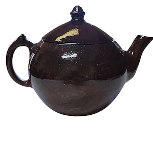 May include: A dark brown ceramic teapot with a matching lid. The teapot has a rounded body, a curved handle, and a spout. The lid is topped with a small knob. The teapot is likely used for brewing tea.