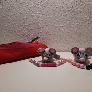 Könnte beinhalten: Zwei gehäkelte Schnullerhalter mit Mäusemotiven, grauen Körpern, rosa Ohren und Nasen sowie Perlenketten. Eine rote Federtasche und Stifte sind im Hintergrund. Die Clips sind handgefertigt und für Babys geeignet.