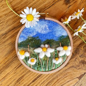 Peut inclure: Un cercle de broderie circulaire avec une scène de paysage en feutre représentant un ciel bleu, de l'herbe verte et des marguerites blanches. Le cercle est sur une surface en bois avec une seule marguerite blanche au centre jaune.