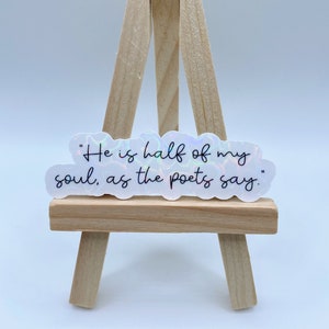 May include: A white sticker with a holographic finish features the quote "He is half of my soul, as the poets say." The sticker is displayed on a small wooden easel.