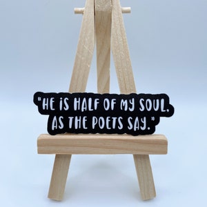May include: A black sticker with white text that reads "HE IS HALF OF MY SOUL. AS THE POETS SAY." The sticker is displayed on a small wooden easel.