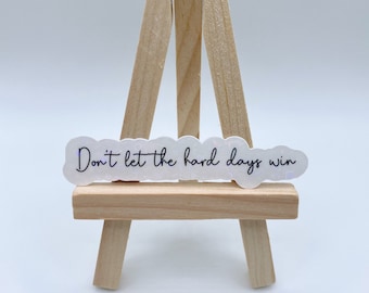 May include: A white sticker with the text "Don't let the hard days win" in black script. The sticker has a cloud-like shape and is displayed on a small wooden easel against a white background. The sticker is approximately 4 inches long.