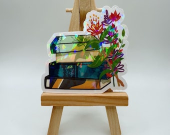 May include: A holographic sticker featuring a stack of books with a floral bouquet. The books have teal, green, and black covers. The flowers are red, purple, and orange with green leaves. The sticker is displayed on a small wooden easel.