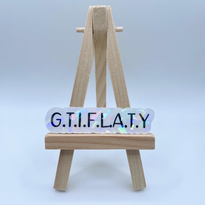May include: A white iridescent sticker with the text "G.T.I.F.L.A.T.Y." in black lettering. The sticker is displayed on a small wooden easel.