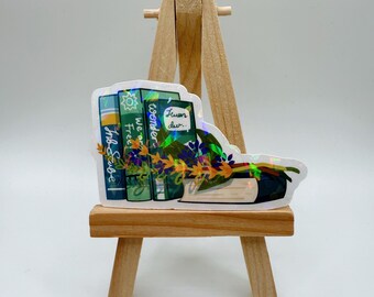 May include: A holographic sticker featuring a stack of books with floral accents. The books have titles like "Ink Scribe," "Free," and "Wonder." The sticker is displayed on a small wooden easel against a white background.