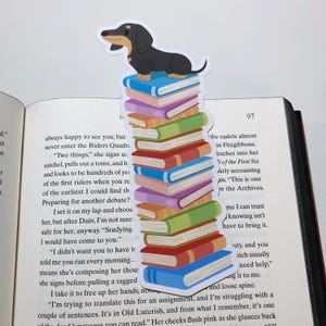 May include: A dachshund bookmark with a black and brown dachshund sitting on top of a stack of colorful books. The bookmark is on a page of a book with text that reads "always happy to see you, but never enter the Riders Quadra. "Two things," she signs as satchel, pulls out a tome, and h and looks to be hundreds of yea of the first riders when you re of the earliest I could find th Preparing for another debate? I set it on my lap and choos her, but after Dain, I'm not sure safe for her, anyway. "Studying. I would have come to you." "I didn't want you to have to told me you run every morning... means she's composing her thou she signs before pulling a ragged I take it to free up her hands, no "I'm trying to translate this for an assignment, and I'm struggling with a couple of sentences. It's in Old Lucerish, and from what I remember, it's one of the dead languages you can read." Her cheeks flush pink as she glances back"