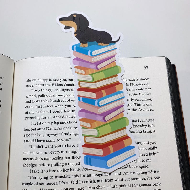 May include: A dachshund bookmark with a black and brown dachshund sitting on top of a stack of colorful books. The bookmark is on a page of a book with text that reads "always happy to see you, but never enter the Riders Quadra. "Two things," she signs as satchel, pulls out a tome, and h and looks to be hundreds of yea of the first riders when you re of the earliest I could find th Preparing for another debate? I set it on my lap and choos her, but after Dain, I'm not sure safe for her, anyway. "Studying. I would have come to you." "I didn't want you to have to told me you run every morning... means she's composing her thou she signs before pulling a ragged I take it to free up her hands, no "I'm trying to translate this for an assignment, and I'm struggling with a couple of sentences. It's in Old Lucerish, and from what I remember, it's one of the dead languages you can read." Her cheeks flush pink as she glances back"
