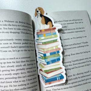 May include: A whimsical bookmark featuring a beagle dog sitting atop a stack of colorful books. The bookmark is white with a die-cut shape, and the books have blue, green, and pink spines. The bookmark is placed in an open book.