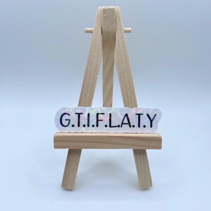 May include: A white iridescent sticker with the text "G.T.I.F.L.A.T.Y." in black lettering. The sticker is displayed on a small wooden easel.