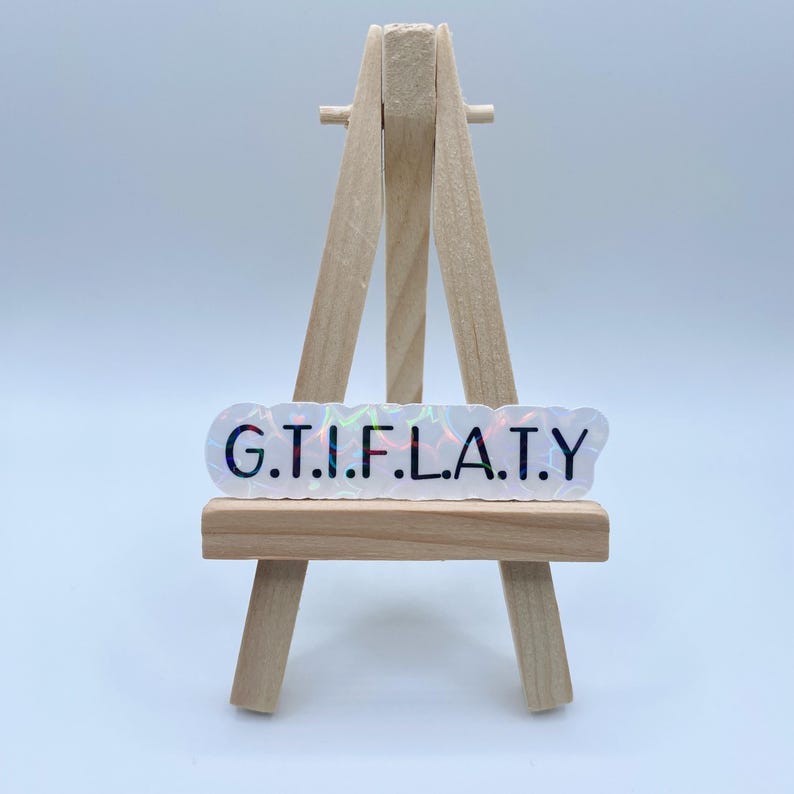 May include: A white iridescent sticker with the text "G.T.I.F.L.A.T.Y." in black lettering. The sticker is displayed on a small wooden easel.