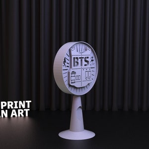 May include: A 3D printed model of the BTS logo, featuring a stylised bulletproof vest with the band's name in bold letters. The model is mounted on a circular base with a cone-shaped stand. The background is a black curtain.