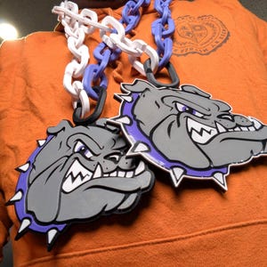 May include: A large, gray bulldog pendant with purple and white accents hangs from a chain. The pendant features a detailed bulldog head with a fierce expression and a purple collar. The chain is a mix of white, blue, and black links.