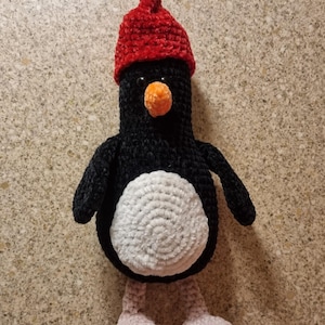 Puede incluir: Un pingüino de crochet negro y blanco con un gorro de crochet rojo. El pingüino tiene una barriga blanca y pies rosas.