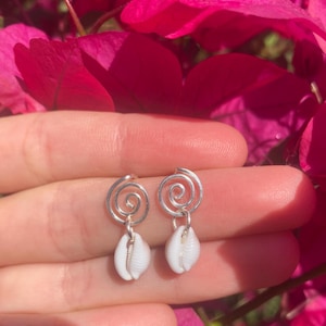 Op de afbeelding: Een paar zilveren spiraalvormige oorbellen met kleine witte schelpen hangers. De oorbellen worden gepresenteerd tegen een achtergrond van felroze bloemen, wat een contrast van kleuren en texturen creëert. Een delicaat sieraad.