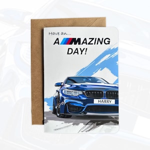 May include: A greetings card featuring a blue sports car illustration with the text Have an... A-MAZING DAY! and a personalised number plate reading HARRY. The card is positioned in front of a brown paper envelope on a light background.