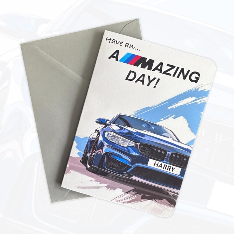 May include: A greeting card with a blue sports car illustration and the text Have an A-MAZING DAY! The car features a license plate reading HARRY and the BMW M logo. A plain grey envelope is positioned behind the card on a white background.