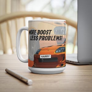 May include: White ceramic mug with an orange sports car graphic and the slogan "MORE BOOST LESS PROBLEMS!". The mug also displays the name "HARRY" on the license plate. It has a large handle and a wide base.