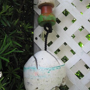 May include: A weathered white fishing buoy with a teal horizontal stripe, suspended by a black rope. Above the main buoy are three smaller, stacked cylindrical floats in beige, orange, and green, showing signs of wear and texture.