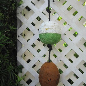 May include: A vertical string of two nautical buoys suspended by a black rope. The top buoy is spherical with a white upper half and a green lower half. The bottom buoy is oval-shaped and brown with a textured surface and a small circular opening.
