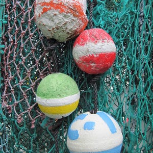 May include: Four spherical fishing floats with weathered paint textures, suspended by black ropes against a green nylon fishing net. The floats feature white, orange, red, green, yellow, and blue color patterns with visible signs of wear.